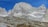 A rustic mountain cabin nestled at the base of towering, rugged granite peaks. The cabin is surrounded by rocky and grassy terrain under a clear blue sky.