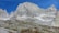 A rustic mountain cabin nestled at the base of towering, rugged granite peaks. The cabin is surrounded by rocky and grassy terrain under a clear blue sky.