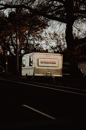 A white RV camper with the brand name 'Windsor' is parked by the side of a road. The scene is surrounded by large, leafless trees that suggest a late autumn or early winter atmosphere. The lighting appears to be low, possibly during twilight, casting deep shadows across the scene. Other vehicles can be faintly seen in the background among the trees.