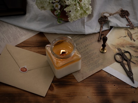A cozy scene featuring a lit candle in a glass jar placed on a wooden surface, surrounded by vintage items including an envelope with a pumpkin sticker, antique scissors, a wax seal stamp, and old papers with printed music and bird illustrations. A bouquet of white flowers is nestled nearby, adding a fresh touch.