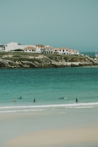 A serene coastal scene featuring several surfers in the turquoise water near a sandy beach. In the background, there are white houses with red-tiled roofs situated on a rocky shoreline.