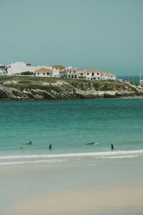 A serene coastal scene featuring several surfers in the turquoise water near a sandy beach. In the background, there are white houses with red-tiled roofs situated on a rocky shoreline.