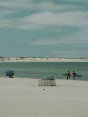 Two surfers with surfboards walk on a sandy beach towards the water. The ocean is a calm blue-green, and the sky is cloudy with a soft grey hue. A couple of umbrellas are set up on the beach, with one featuring a colorful striped pattern, adding a touch of vibrancy to the scene.