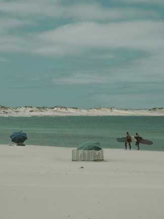 Two surfers with surfboards walk on a sandy beach towards the water. The ocean is a calm blue-green, and the sky is cloudy with a soft grey hue. A couple of umbrellas are set up on the beach, with one featuring a colorful striped pattern, adding a touch of vibrancy to the scene.