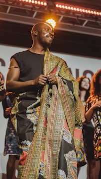 A person wearing a colorful, patterned garment stands on a stage under bright lights. They have face paint and are holding the fabric, smiling with a confident expression. Several people in vibrant clothing stand in the background, suggesting a cultural or fashion event.