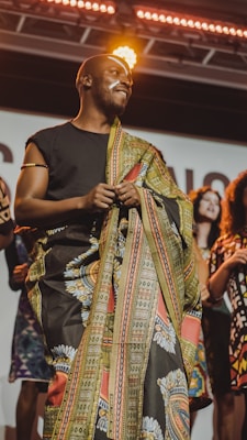 A person wearing a colorful, patterned garment stands on a stage under bright lights. They have face paint and are holding the fabric, smiling with a confident expression. Several people in vibrant clothing stand in the background, suggesting a cultural or fashion event.