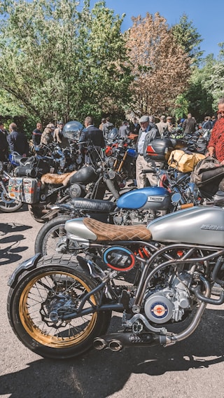 A group of people gathered around various motorcycles in an outdoor setting with lush trees in the background. The motorcycles, including both modern and vintage styles, are parked close together. People are dressed casually, some wearing helmets and gear.
