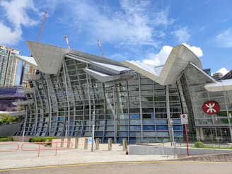 A modern architectural structure featuring a large glass façade with metal accents, set against a clear blue sky. The building has a unique, angular design and includes construction cranes in the background. There is a red symbol on the side of the building, possibly indicating a transportation hub.