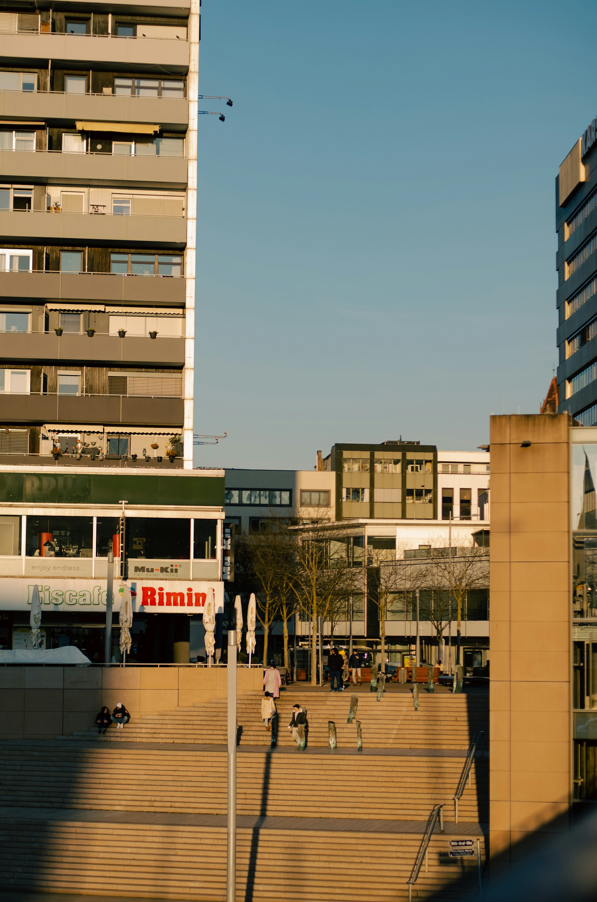 Urban scene with modern multi-story buildings in the background. At the center, a wide staircase leads up, with a few people walking or sitting. Trees line the edge of the stairs, and there are cafes and shops at street level. The sky is clear, and sunlight casts long shadows across the scene.