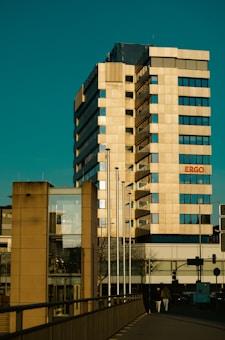 A tall, modern office building with a beige facade, rectangular windows, and multiple stories. The structure features an attached smaller building with large glass panels. Several flagpoles are in front, and two people are walking along a pathway at the building's base. The name 'ERGO' is visible on the building.