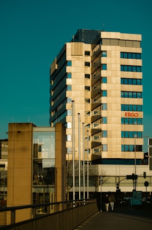 A tall, modern office building with a beige facade, rectangular windows, and multiple stories. The structure features an attached smaller building with large glass panels. Several flagpoles are in front, and two people are walking along a pathway at the building's base. The name 'ERGO' is visible on the building.