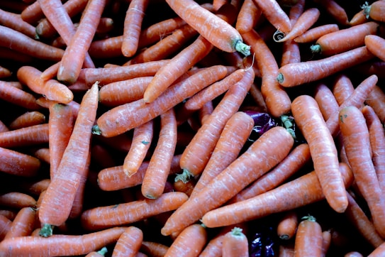 A large pile of fresh, orange carrots with green stems, closely packed together. The carrots vary slightly in size and shape, and a few purple vegetables are visible among them.