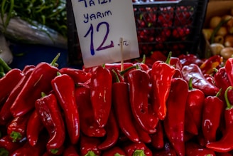 A vibrant display of red peppers stacked on top of each other with a handwritten sign indicating a price. The background reveals part of a market setting with other vegetables.