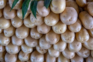 A close-up view of a large pile of smooth, light brown potatoes, stacked in an orderly manner. A few green leaves protrude from the top left corner, adding a touch of color contrast.