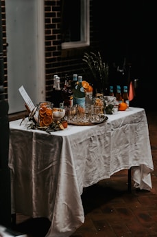 A table covered with a white tablecloth is set up with various bottles and glassware. There are several bottles of beverages, a few glasses, and some garnishes like slices of orange. The setting gives a rustic and inviting feel.