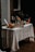 A table covered with a white tablecloth is set up with various bottles and glassware. There are several bottles of beverages, a few glasses, and some garnishes like slices of orange. The setting gives a rustic and inviting feel.