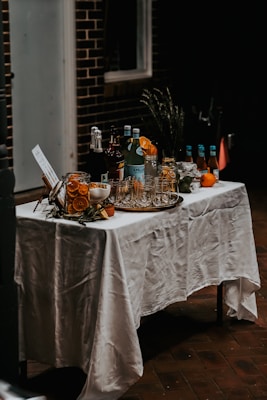 A table covered with a white tablecloth is set up with various bottles and glassware. There are several bottles of beverages, a few glasses, and some garnishes like slices of orange. The setting gives a rustic and inviting feel.