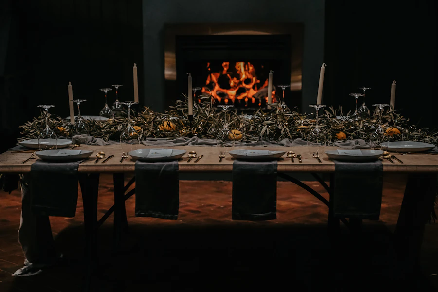 A long wooden table set for dinner beside a warm fireplace glow