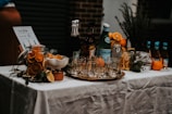 A beautifully arranged cocktail bar setup on a table covered with a white tablecloth. There are various bottles of liquor and sparkling water, along with jars filled with sliced oranges and other citrus fruits. Glasses are neatly arranged on a metallic tray, and an assortment of fresh oranges, lemons, and limes are placed around. A small menu or sign is displayed on the left.