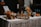 A beautifully arranged cocktail bar setup on a table covered with a white tablecloth. There are various bottles of liquor and sparkling water, along with jars filled with sliced oranges and other citrus fruits. Glasses are neatly arranged on a metallic tray, and an assortment of fresh oranges, lemons, and limes are placed around. A small menu or sign is displayed on the left.