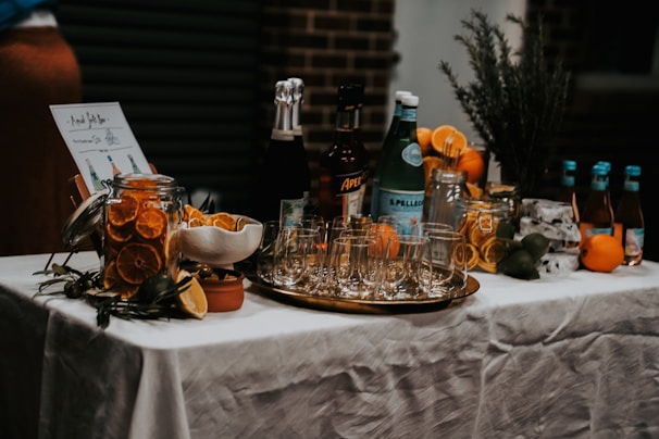 A beautifully arranged cocktail bar setup on a table covered with a white tablecloth. There are various bottles of liquor and sparkling water, along with jars filled with sliced oranges and other citrus fruits. Glasses are neatly arranged on a metallic tray, and an assortment of fresh oranges, lemons, and limes are placed around. A small menu or sign is displayed on the left.