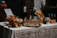A beautifully arranged cocktail bar setup on a table covered with a white tablecloth. There are various bottles of liquor and sparkling water, along with jars filled with sliced oranges and other citrus fruits. Glasses are neatly arranged on a metallic tray, and an assortment of fresh oranges, lemons, and limes are placed around. A small menu or sign is displayed on the left.