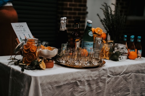 A beautifully arranged cocktail bar setup on a table covered with a white tablecloth. There are various bottles of liquor and sparkling water, along with jars filled with sliced oranges and other citrus fruits. Glasses are neatly arranged on a metallic tray, and an assortment of fresh oranges, lemons, and limes are placed around. A small menu or sign is displayed on the left.