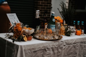 A beautifully arranged cocktail bar setup on a table covered with a white tablecloth. There are various bottles of liquor and sparkling water, along with jars filled with sliced oranges and other citrus fruits. Glasses are neatly arranged on a metallic tray, and an assortment of fresh oranges, lemons, and limes are placed around. A small menu or sign is displayed on the left.