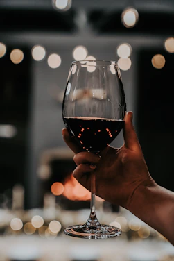 A close-up of a hand gently holding a crystal wine glass filled with deep red wine, with a blurred background of oak barrels.