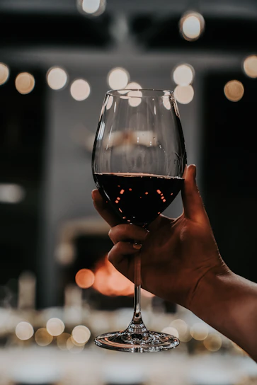 A close-up of a hand gently holding a crystal wine glass filled with deep red wine, with a blurred background of oak barrels.