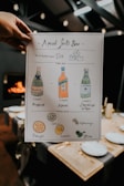 A hand holds a stylishly designed recipe card titled 'Aperol Spritz Bar'. The card illustrates the drink-making process with drawings and text. It includes bottles labeled as Bubbly, Aperol, and Sparkling Water, along with citrus garnishes like orange and lemon, and other garnishes like lime, rosemary, and olive. The background shows a dimly lit dining area with table settings and a warm fire.