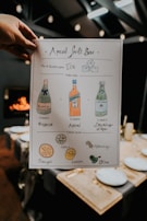 A hand holds a stylishly designed recipe card titled 'Aperol Spritz Bar'. The card illustrates the drink-making process with drawings and text. It includes bottles labeled as Bubbly, Aperol, and Sparkling Water, along with citrus garnishes like orange and lemon, and other garnishes like lime, rosemary, and olive. The background shows a dimly lit dining area with table settings and a warm fire.