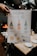 A hand holds a stylishly designed recipe card titled 'Aperol Spritz Bar'. The card illustrates the drink-making process with drawings and text. It includes bottles labeled as Bubbly, Aperol, and Sparkling Water, along with citrus garnishes like orange and lemon, and other garnishes like lime, rosemary, and olive. The background shows a dimly lit dining area with table settings and a warm fire.