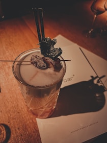 A beautifully crafted cocktail sits on a wooden table, garnished with herbs and a dusting of powdered sugar. The drink is in a textured glass and accompanied by a menu in the background. Warm, ambient lighting casts soft shadows.