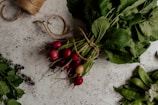 A bundle of freshly picked radishes with green leaves is tied together with twine on a rustic stone surface. To the side, there are some leafy herbs and a bunch of snap peas, creating a farm-fresh and organic vibe.