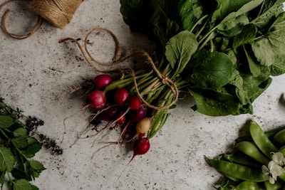 A bundle of freshly picked radishes with green leaves is tied together with twine on a rustic stone surface. To the side, there are some leafy herbs and a bunch of snap peas, creating a farm-fresh and organic vibe.