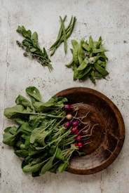 A rustic arrangement of fresh vegetables on a textured surface, featuring leafy radishes, green beans, snow peas, and herbs. The produce is artfully placed around a wooden bowl at the bottom right.