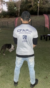 A person stands on a grassy field, wearing a cap, jeans, and a shirt with text related to a training course. In the background, there are two flags and trees. A dog is present on the ground, appearing to be eating or sniffing something.