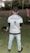 A person stands on a grassy field, wearing a cap, jeans, and a shirt with text related to a training course. In the background, there are two flags and trees. A dog is present on the ground, appearing to be eating or sniffing something.