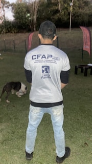 A person stands on a grassy field, wearing a cap, jeans, and a shirt with text related to a training course. In the background, there are two flags and trees. A dog is present on the ground, appearing to be eating or sniffing something.