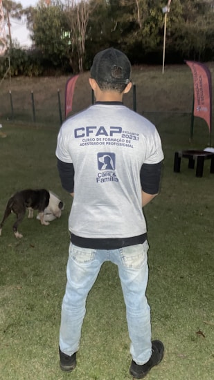 A person stands on a grassy field, wearing a cap, jeans, and a shirt with text related to a training course. In the background, there are two flags and trees. A dog is present on the ground, appearing to be eating or sniffing something.