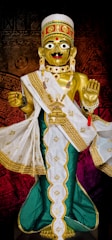 A golden statue adorned with elaborate jewelry and a detailed headdress, wearing a white and green robe with gold embroidery. The figure has large eyes and a red bindi on its forehead, with one hand raised in blessing.