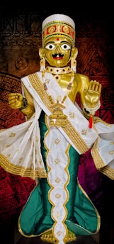 A golden statue adorned with elaborate jewelry and a detailed headdress, wearing a white and green robe with gold embroidery. The figure has large eyes and a red bindi on its forehead, with one hand raised in blessing.