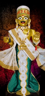 A golden statue adorned with elaborate jewelry and a detailed headdress, wearing a white and green robe with gold embroidery. The figure has large eyes and a red bindi on its forehead, with one hand raised in blessing.
