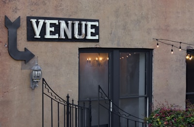 An exterior wall with a large sign that reads 'VENUE' in bold letters above a double-door entrance. An arrow points toward the venue, accompanied by a traditional outdoor wall lantern. A string of decorative lights is hanging above from left to right, adding a cozy and inviting ambiance. Small plants are visible at the bottom of the image, and a decorative metal gate partially obscures the entrance.