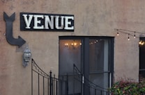 An exterior wall with a large sign that reads 'VENUE' in bold letters above a double-door entrance. An arrow points toward the venue, accompanied by a traditional outdoor wall lantern. A string of decorative lights is hanging above from left to right, adding a cozy and inviting ambiance. Small plants are visible at the bottom of the image, and a decorative metal gate partially obscures the entrance.