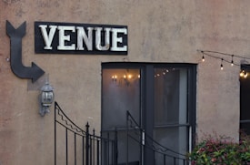 An exterior wall with a large sign that reads 'VENUE' in bold letters above a double-door entrance. An arrow points toward the venue, accompanied by a traditional outdoor wall lantern. A string of decorative lights is hanging above from left to right, adding a cozy and inviting ambiance. Small plants are visible at the bottom of the image, and a decorative metal gate partially obscures the entrance.