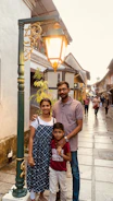 A family of three is standing on a cobblestone street under a vintage street lamp. The street is lined with buildings that have a traditional architectural style, and a few people are walking in the background.