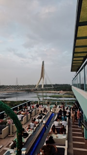 An outdoor rooftop dining area is bustling with people seated at tables, enjoying food and drinks. The setting is modern and vibrant, featuring decorative plants and overhead fans. In the background, a large suspension bridge spans over a river, surrounded by greenery and a partly cloudy sky.