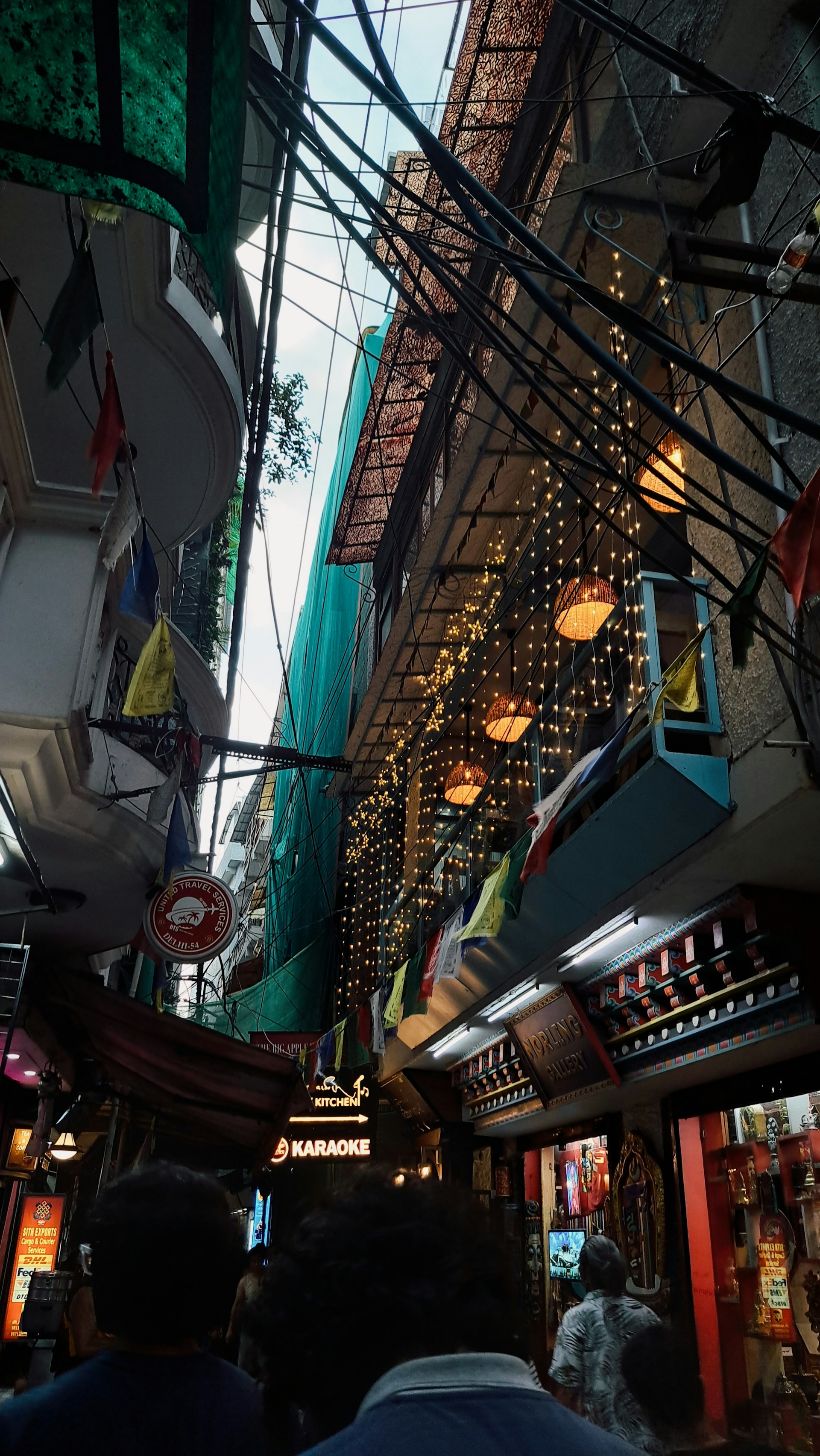 A narrow street crowded with signs, lights, and hanging cables. Colorful flags and decorative lights add vibrancy to the scene, while signage for karaoke and other businesses suggest a lively area.